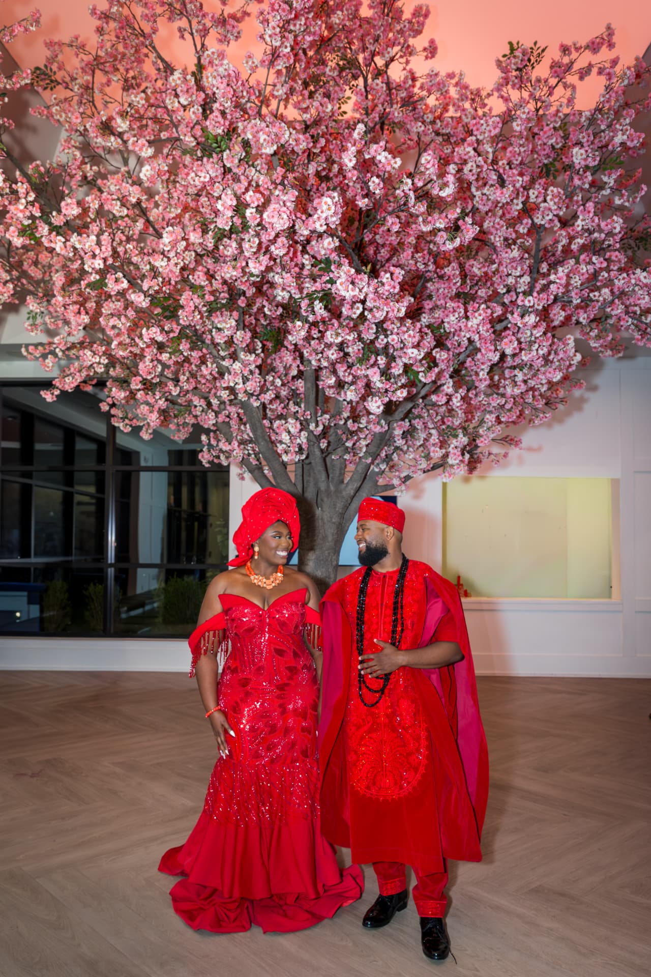 A couple in vibrant red traditional wedding attire beneath a blossoming tree at their reception